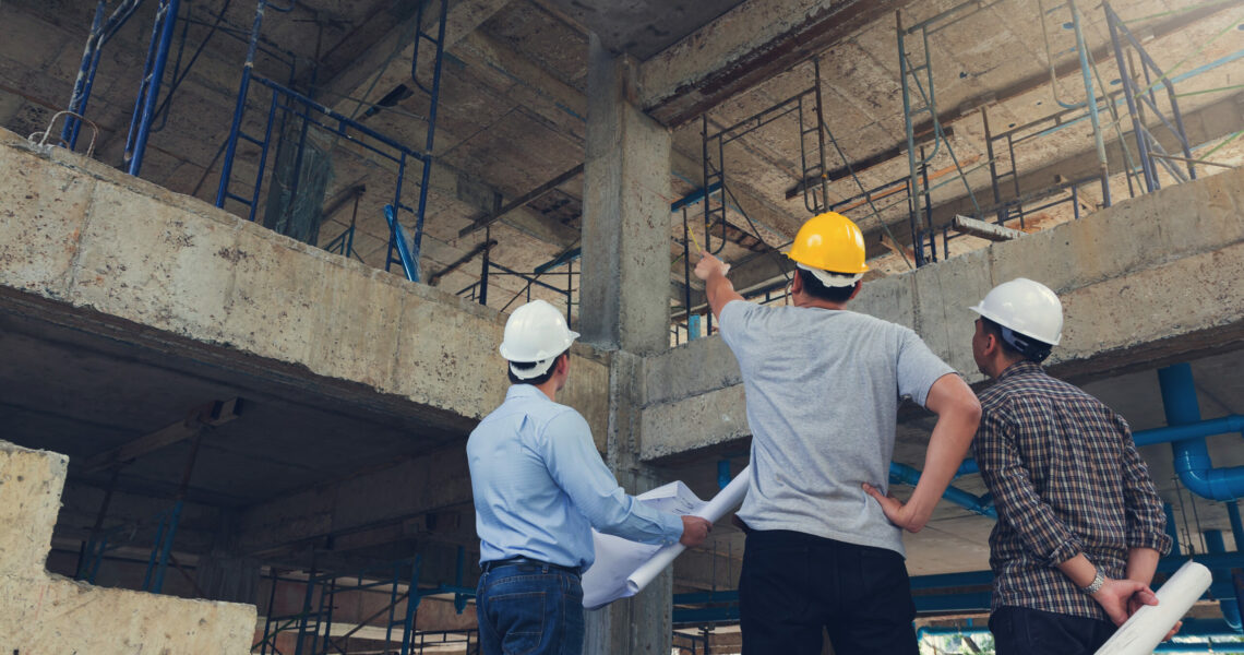 Three construction workers at a job site one pointing up the other holding blueprints
